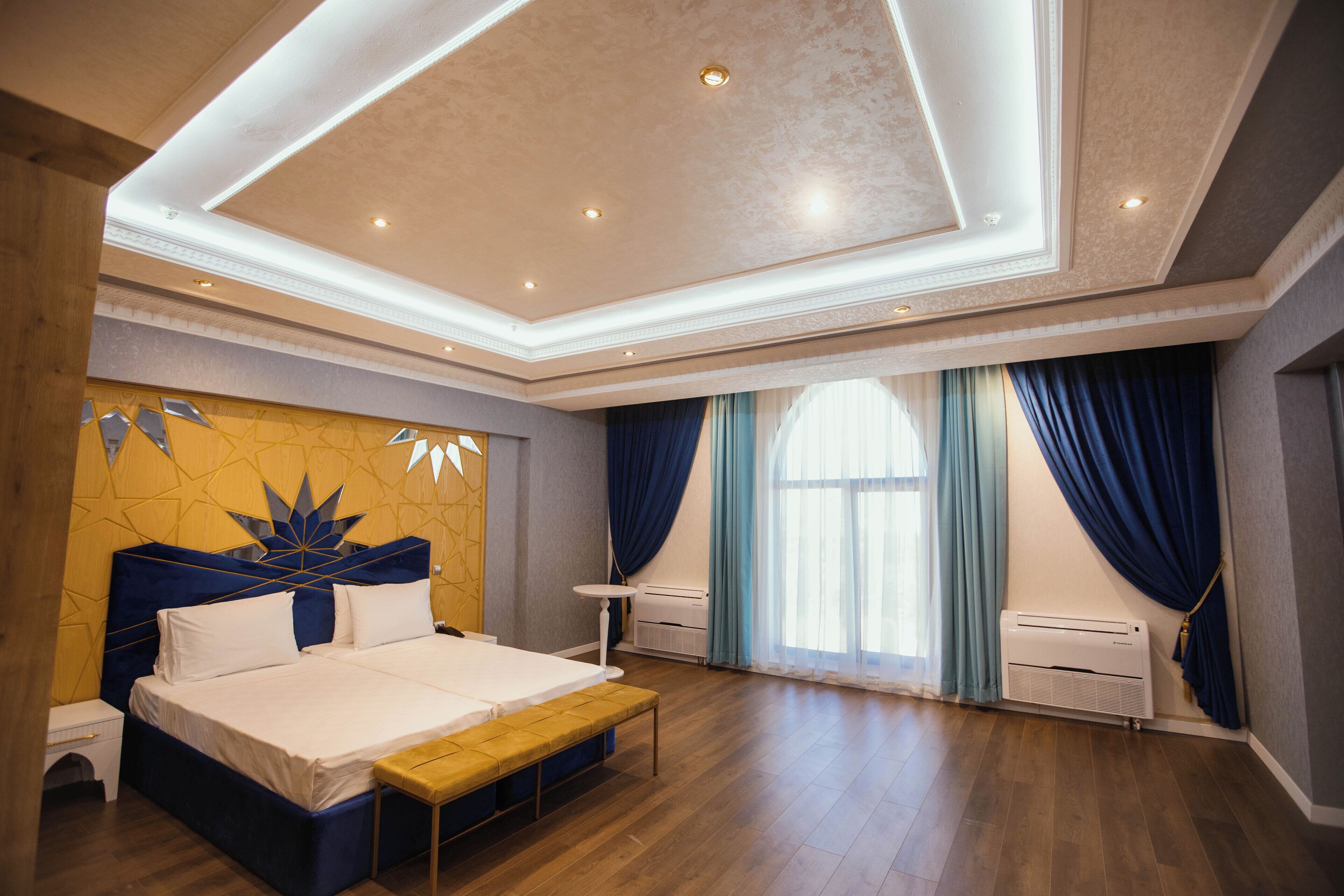 A spacious bedroom with a large bed, a bench, a bedside table, and a window with curtains.