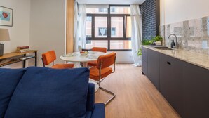 Ein modernes Wohnzimmer mit einem blauen Sofa, einem Esstisch mit orangefarbenen Stühlen, einer Küche mit schwarzem Spritzschutz und einem großen Fenster mit Vorhängen.