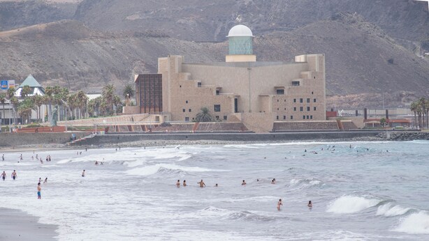 Ein Küstengebäude mit Kuppel und Turm, umgeben von einem Strand mit badenden und surfenden Menschen.