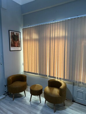 A room with two brown armchairs and a small ottoman, a framed picture on the wall, and a large window with vertical blinds. - KAYNARCA HOTEL (Iznik)