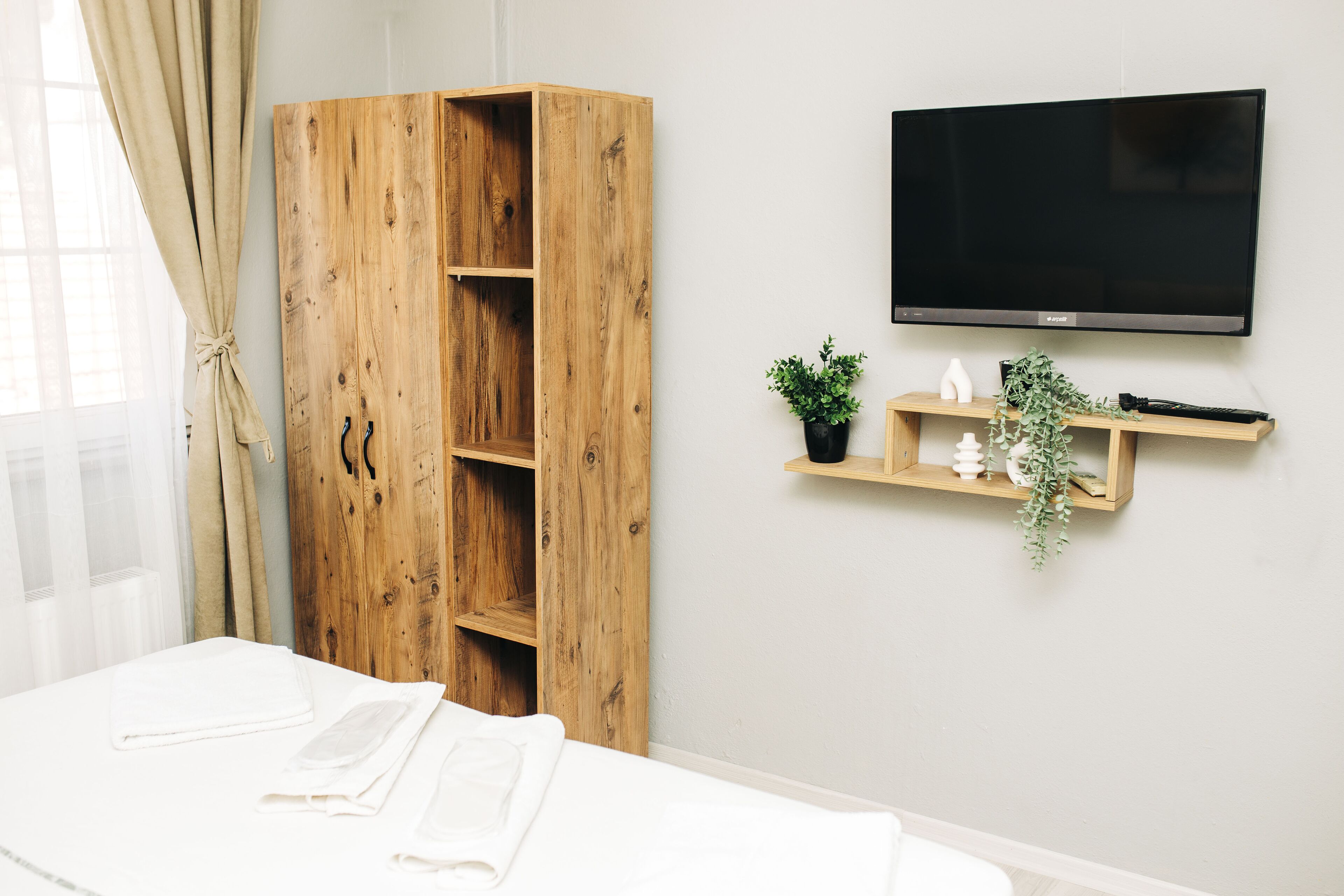 A bedroom with a wooden wardrobe, a mounted TV, and a bedside table with a plant.