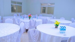 A room with multiple round tables covered with white tablecloths, chairs with white covers, and small potted plants. - Elanacha Hotel (Arusha)