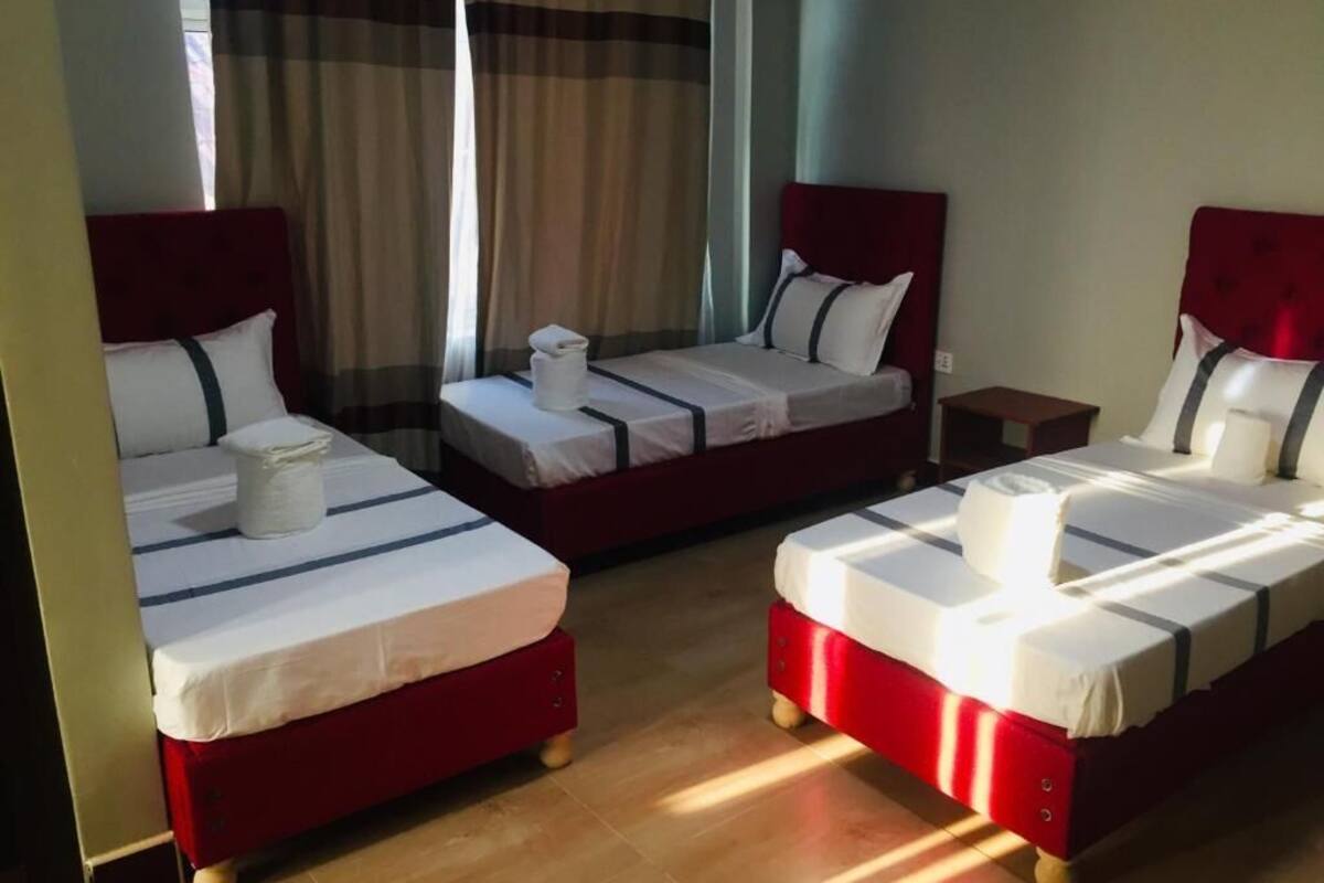 A hotel room with three single beds, each with a red headboard and white bedding. There is a small wooden nightstand between the beds.