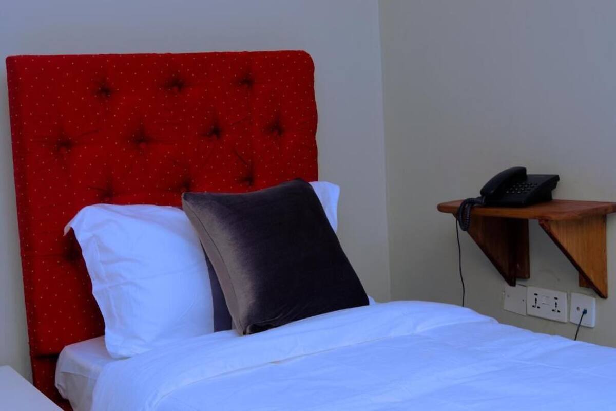 A hotel room with a red upholstered headboard, white bedding, and a bedside telephone on a wooden shelf.