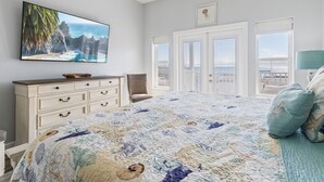 Ein Schlafzimmer mit einem großen Bett, einem an der Wand montierten Fernseher, einem Schminktisch und Blick auf den Ozean durch die Fenster.