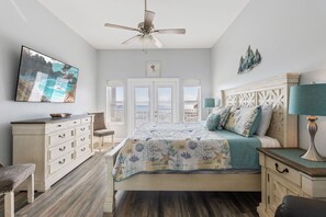 A bedroom with a bed, a dresser, a chair, a ceiling fan, and a TV on the wall.