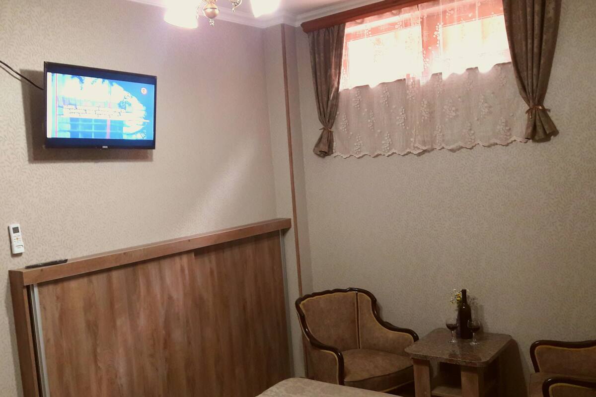 A hotel room with a television mounted on the wall, a wooden headboard, two upholstered chairs, a small table with a bottle and glass, and a window with curtains.