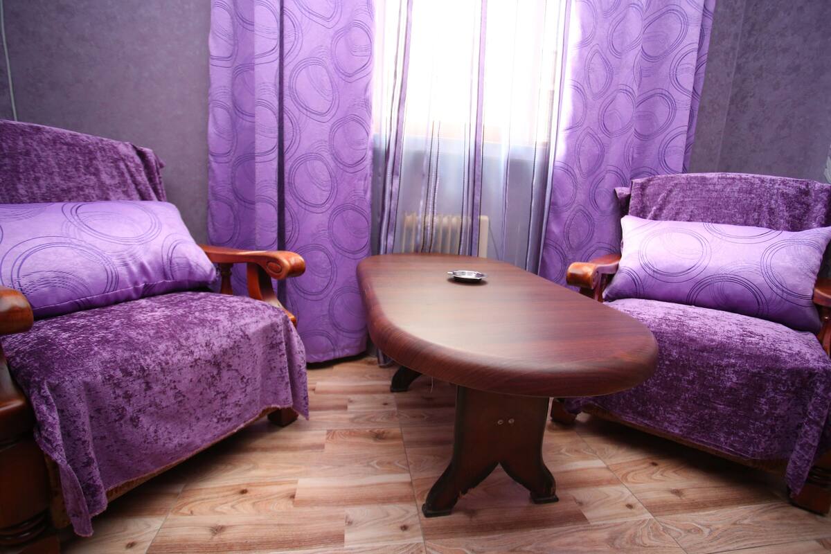 A room with two purple upholstered chairs, a wooden table, and a patterned curtain.