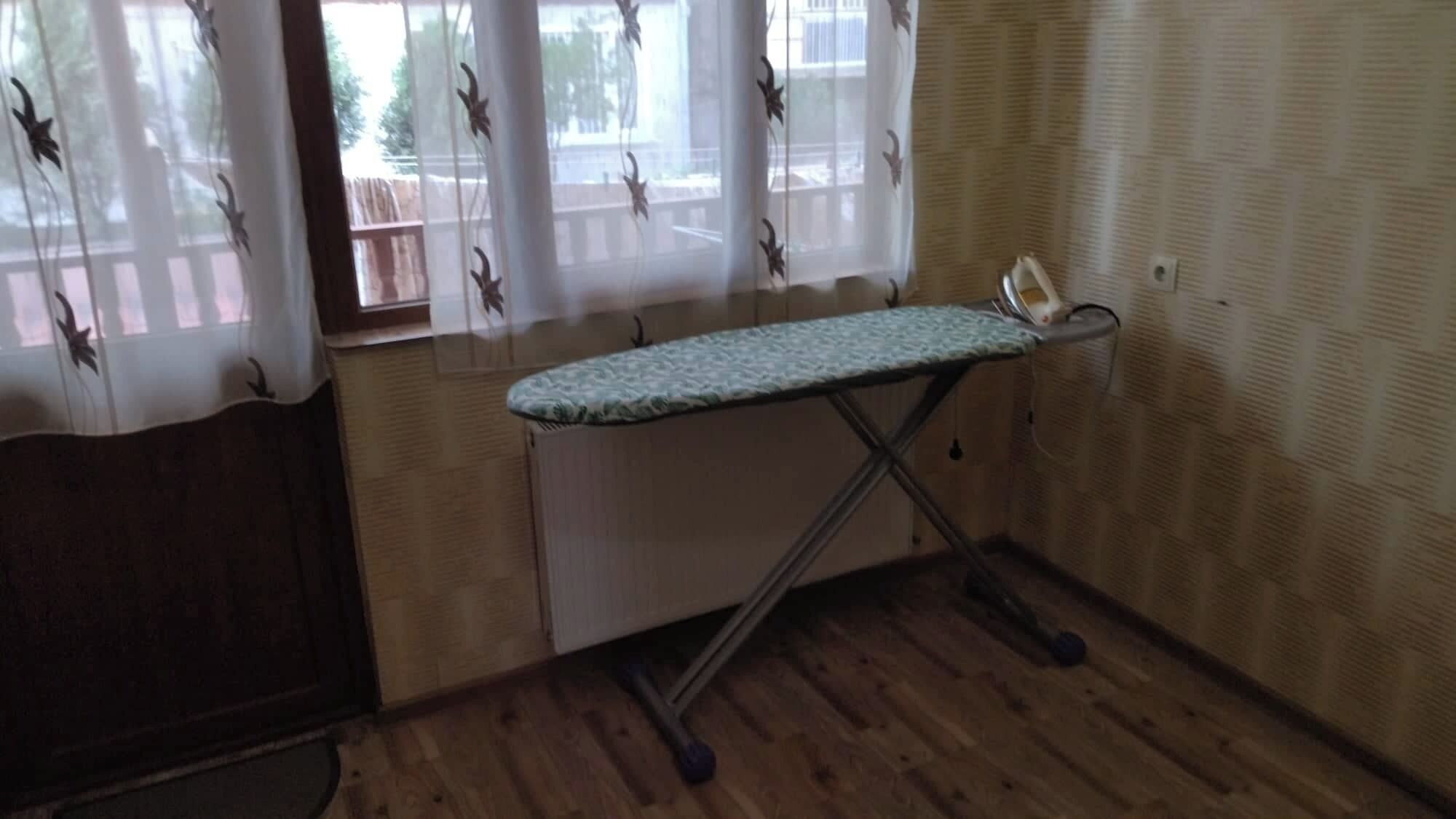 A room with a wooden floor, a window with sheer curtains, and an ironing board with an iron.