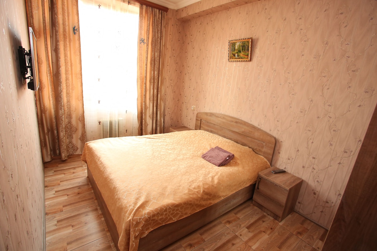 A bedroom with a wooden bed, a nightstand, a painting on the wall, and a window with curtains.