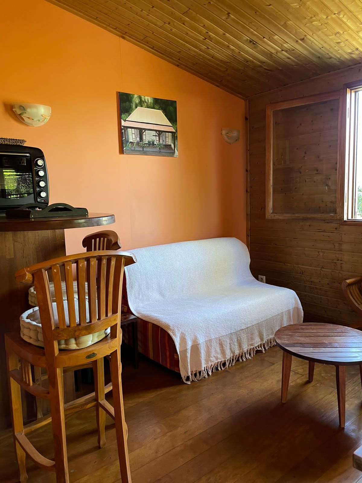 A cozy room with a wooden ceiling, a sofa with a white blanket, a wooden chair, a small table, and a picture on the wall.