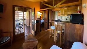 L’intérieur chaleureux d’un chalet avec un plafond en bois, un coin repas comprenant une table et des chaises, une cuisine avec des meubles en bois et une porte-fenêtre coulissante donnant sur une terrasse extérieure.