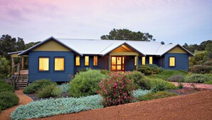 Une maison à deux étages, dotée d’un toit à pignon, de grandes fenêtres et d’un jardin bien entretenu où poussent différentes espèces de plantes et un petit arbre.