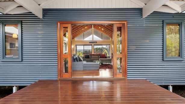 Une maison moderne dotée d’une grande terrasse en bois, d’un espace de vie spacieux avec un canapé et une table basse, et offrant une vue sur l’extérieur grâce à de grandes fenêtres.