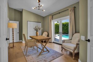 Ein Zimmer mit einem Holztisch, zwei Stühlen, einer Blumenvase, einer Lampe und Blick auf einen Swimmingpool durch eine Glastür.