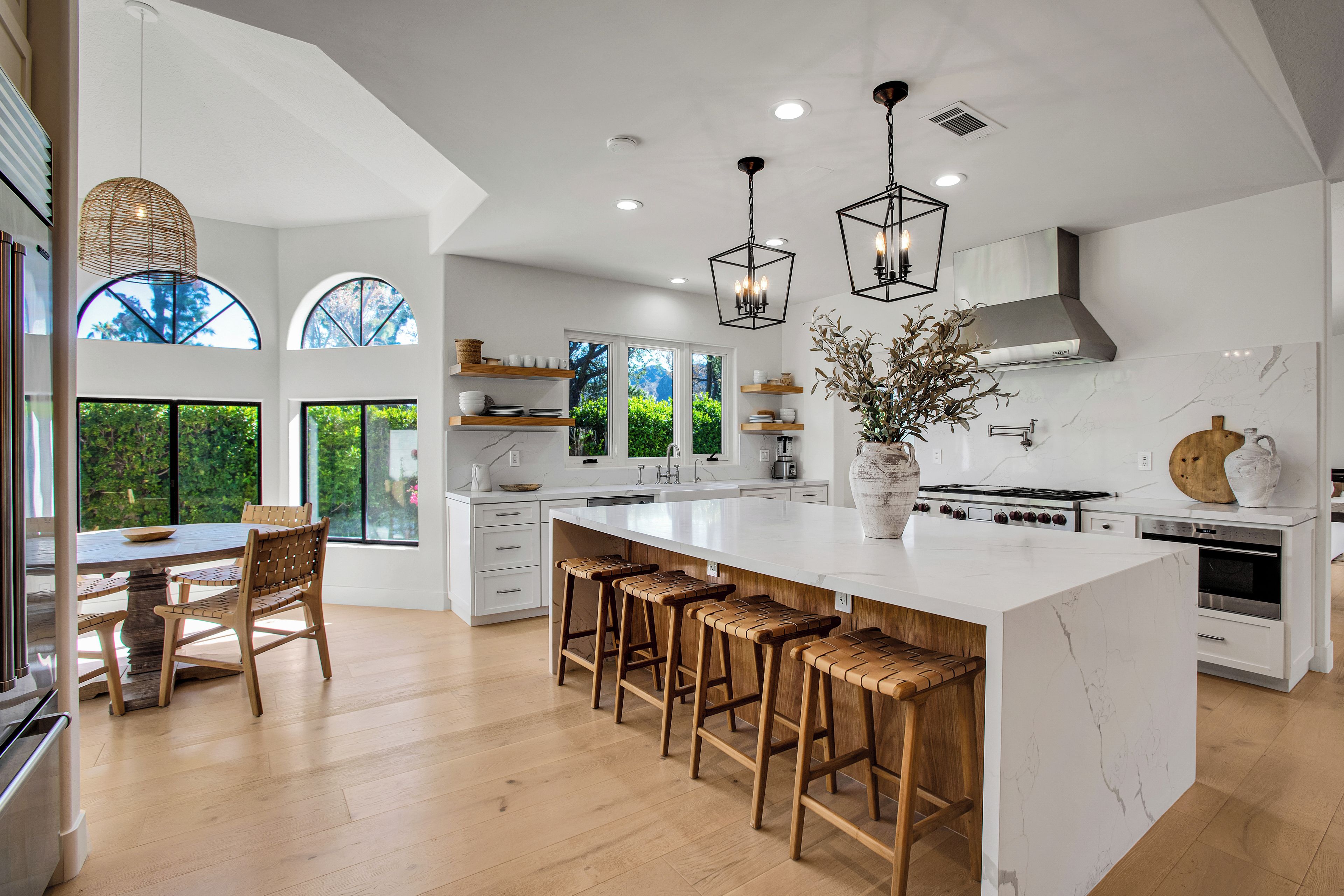 A modern kitchen with a large island, wooden bar stools, and a view of greenery through the windows.
