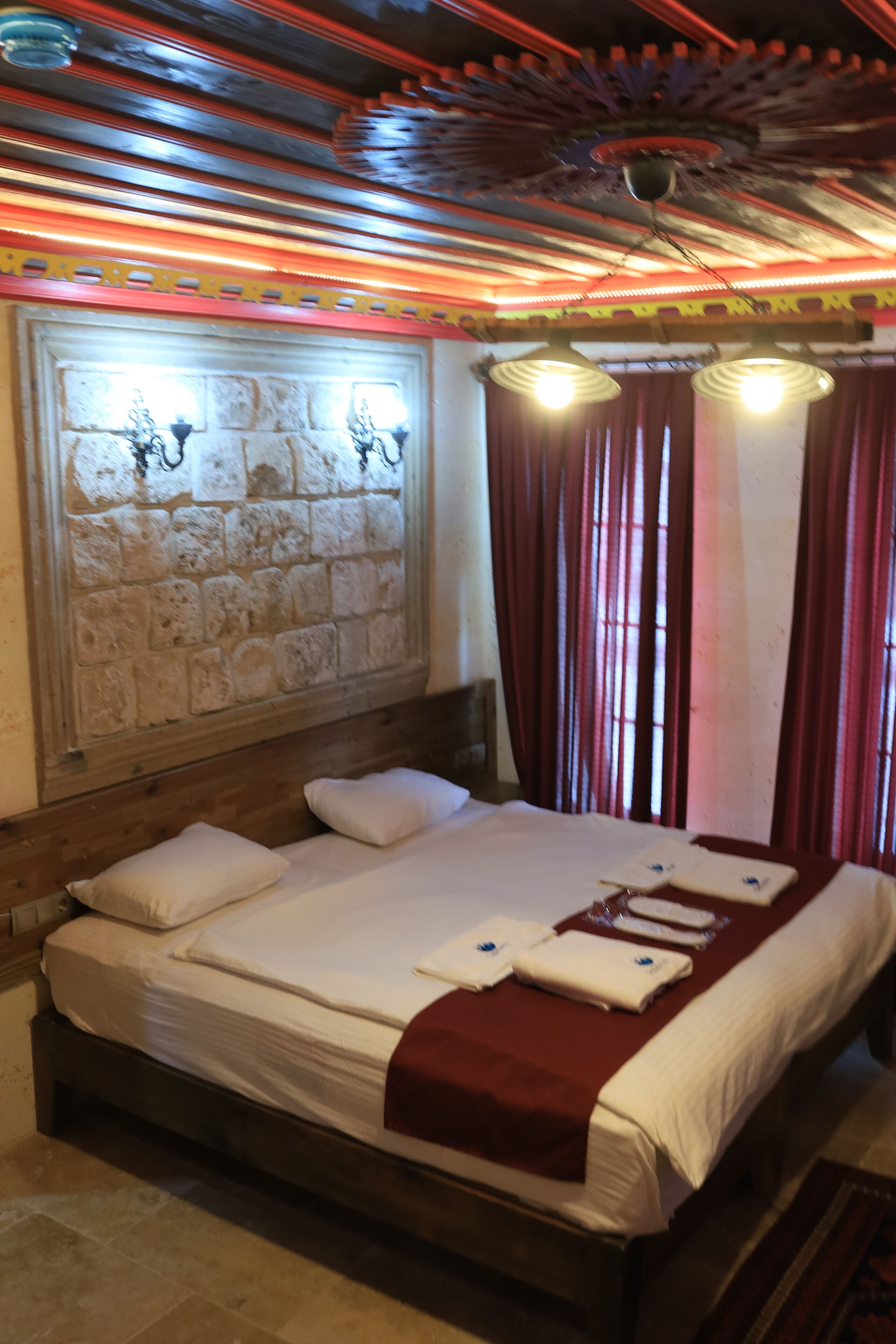 A neatly made bed with white linens and a red blanket, a stone wall with a decorative pattern, and a ceiling with a unique design.