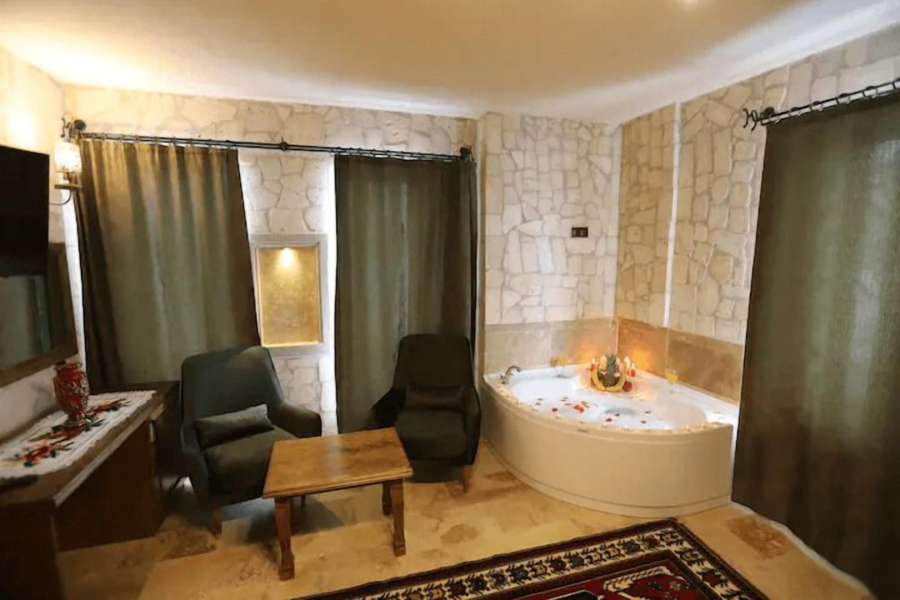 A room with a stone wall, a large bathtub with rose petals, two chairs, a small table, and a curtain.