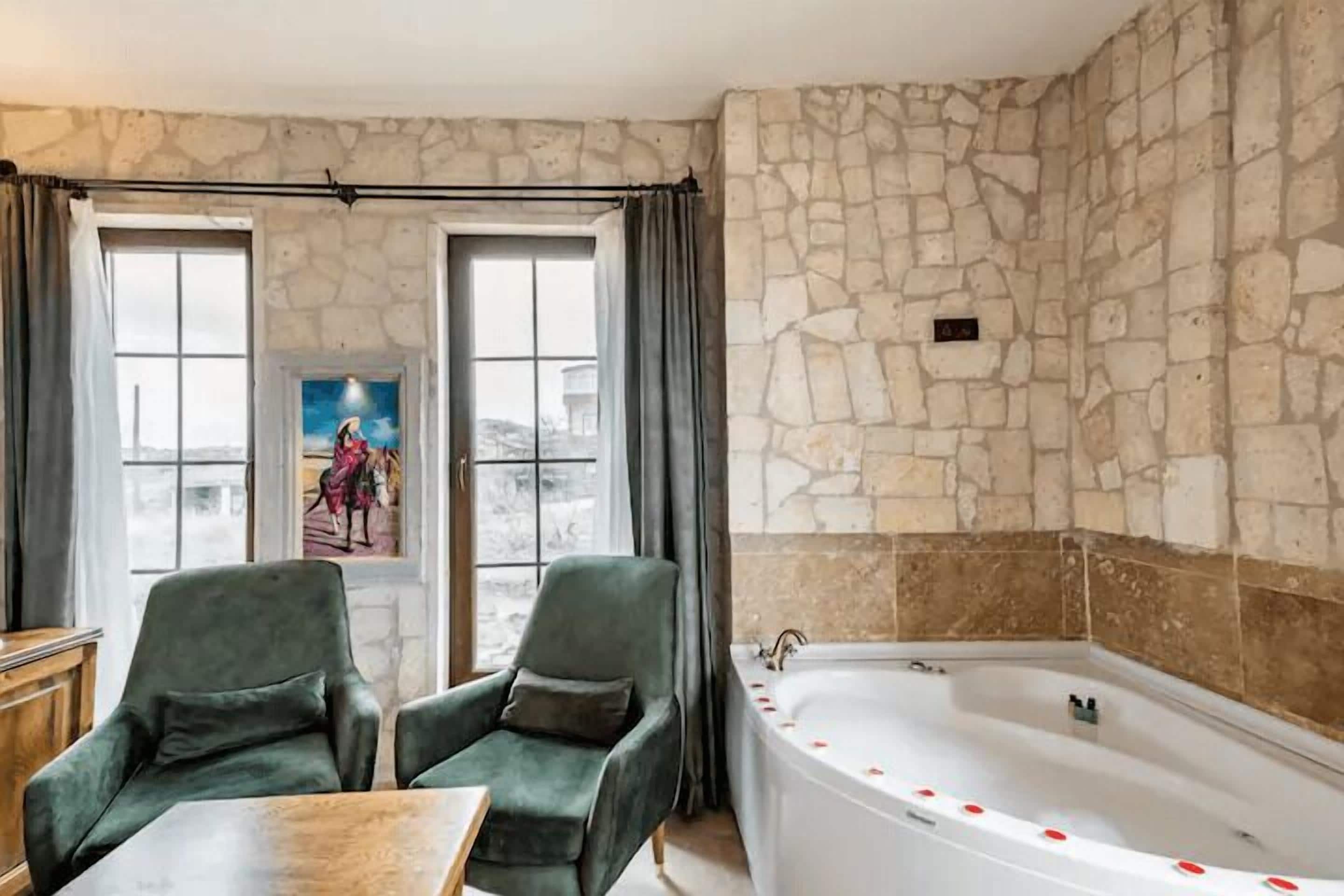 A room with a stone wall, a bathtub, two green armchairs, and a table.