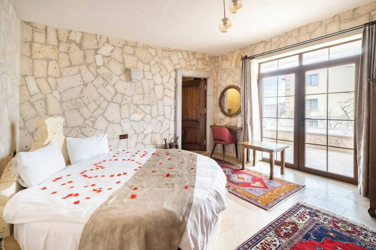 A bedroom with a stone wall, a bed with rose petals, a red chair, a wooden table, and a view through large windows.