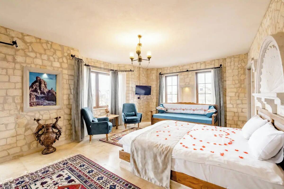 A bedroom with a stone wall, a bed with rose petals, two armchairs, a TV, and a chandelier.