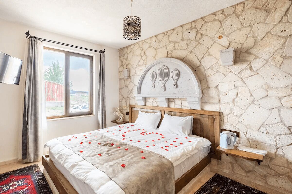 A bedroom with a stone wall, a bed with a white bedspread, a wooden headboard, a bedside table with a kettle, and a TV mounted on the wall.