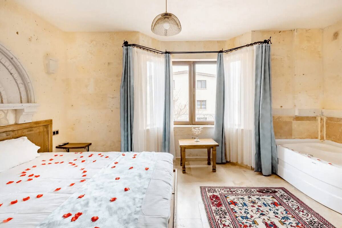 A bedroom with a bed, a small table, a chair, a window with curtains, and a bathtub.