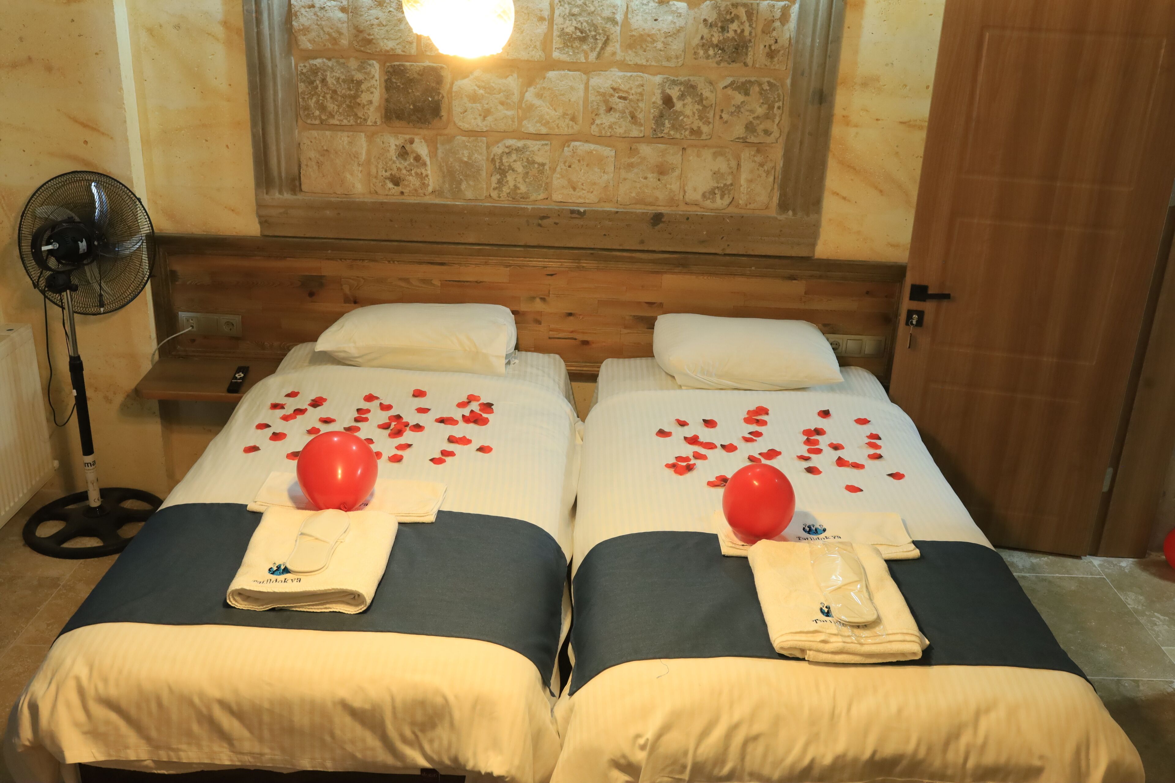 A room with two beds, each with a pillow, a towel, and a red balloon. There is a fan and a stone wall in the background.