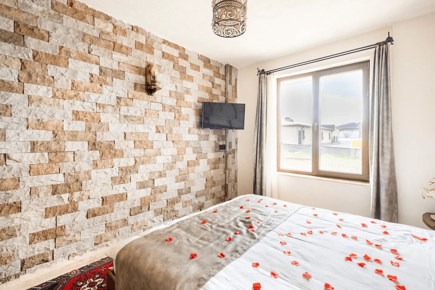 A bedroom with a brick wall, a flat-screen TV, a large window with curtains, and a bed with rose petals.