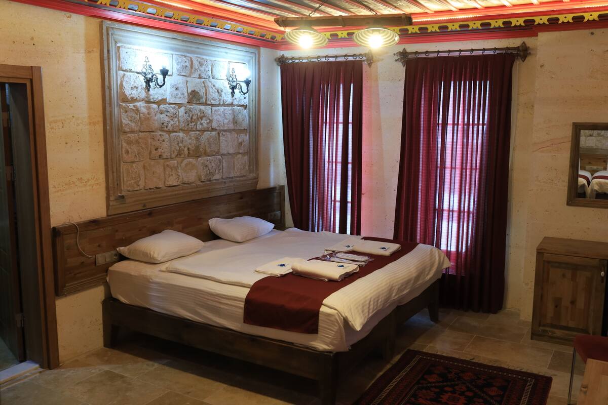 A neatly made bed with white linens and a red blanket, a stone wall with a light fixture, and a wooden cabinet.