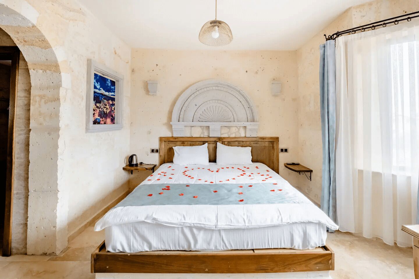 A bedroom with a wooden bed, a white headboard, a decorative wall arch, a small bedside table with a lamp, and a window with sheer curtains.