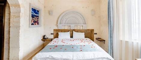 A bedroom with a wooden bed, a white headboard, a decorative wall arch, a small bedside table with a lamp, and a window with sheer curtains.