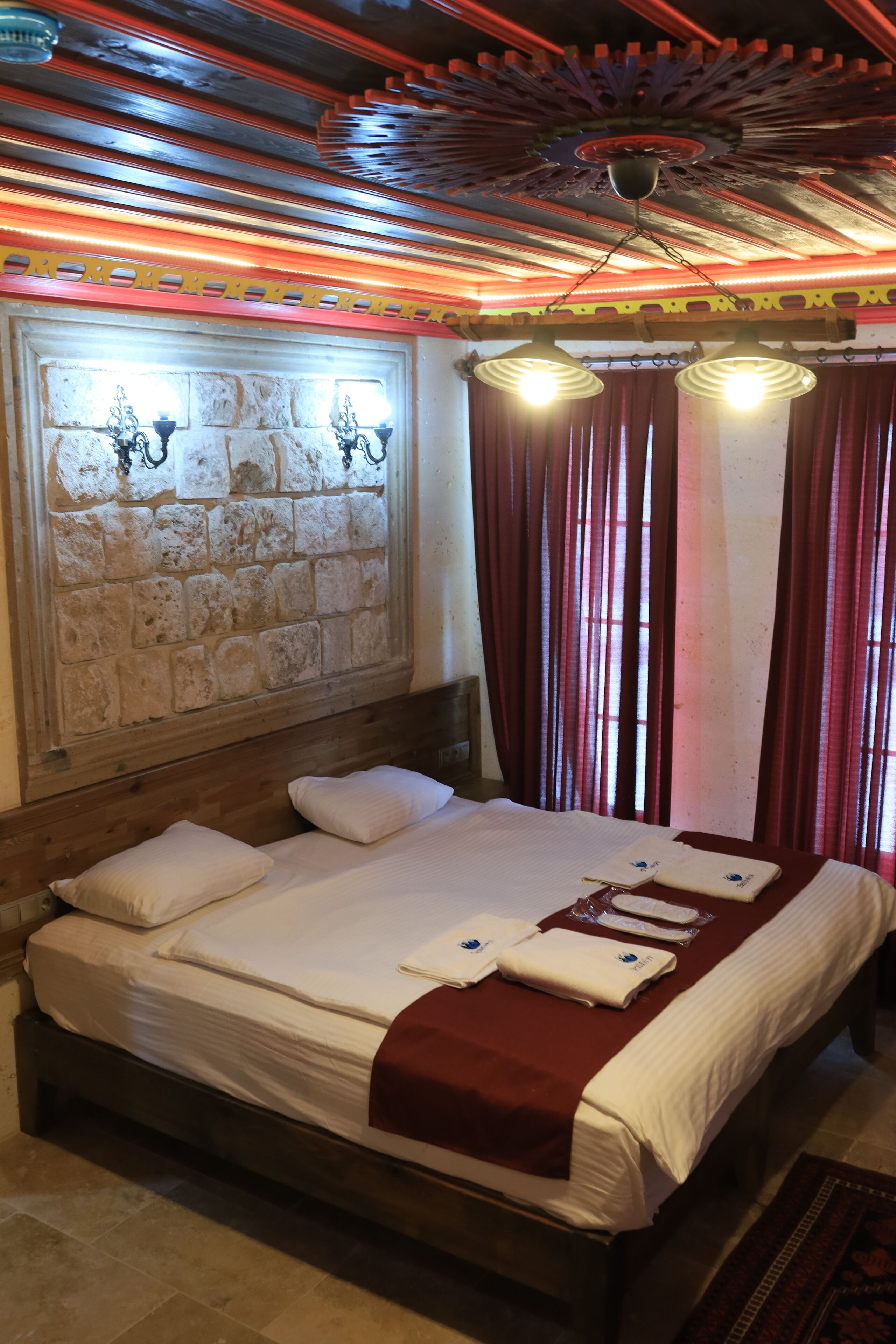 A neatly made bed with a red blanket, a stone wall with a light fixture, and red curtains.