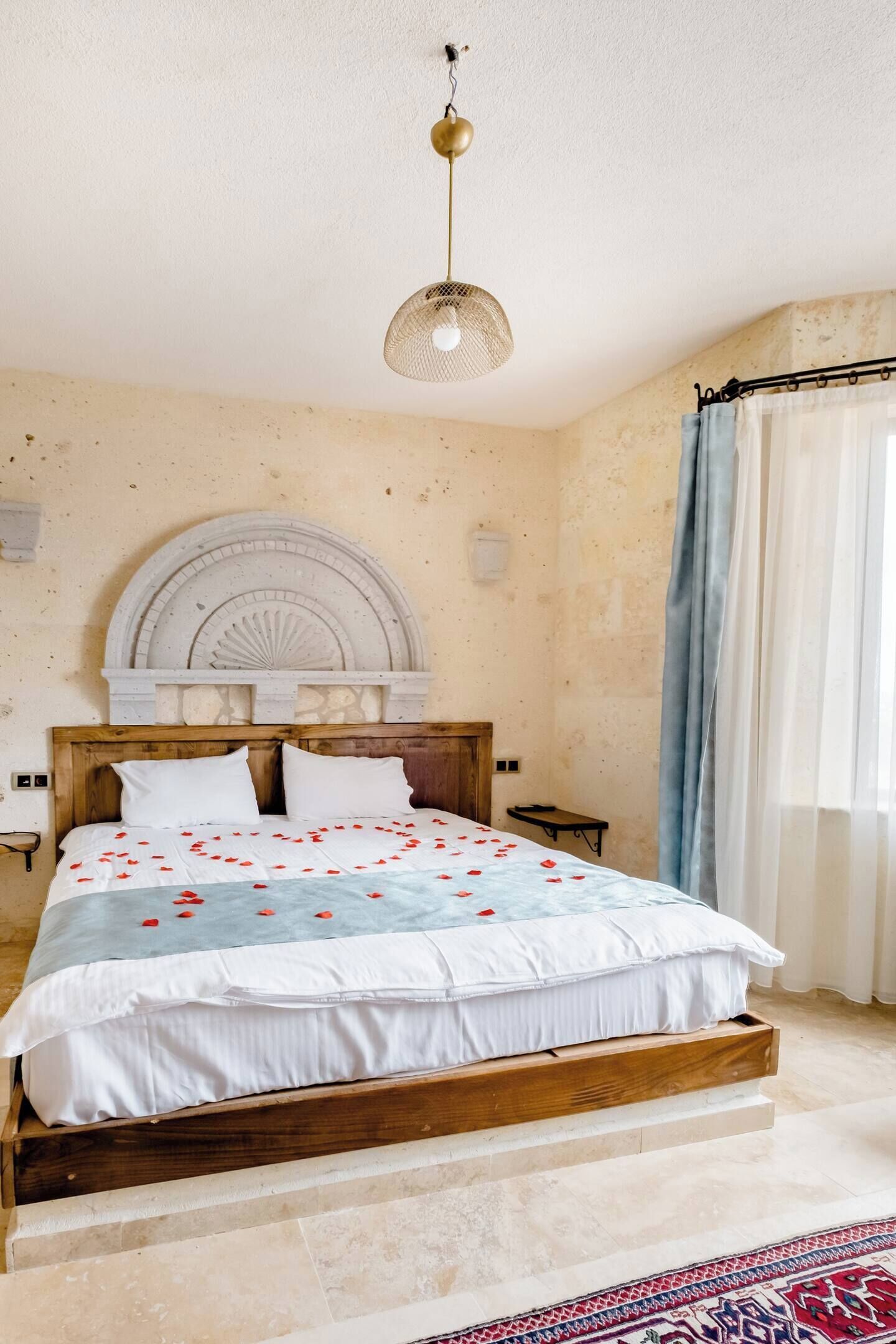 A bedroom with a wooden bed, a decorative ceiling light, a stone wall, and a window with curtains.
