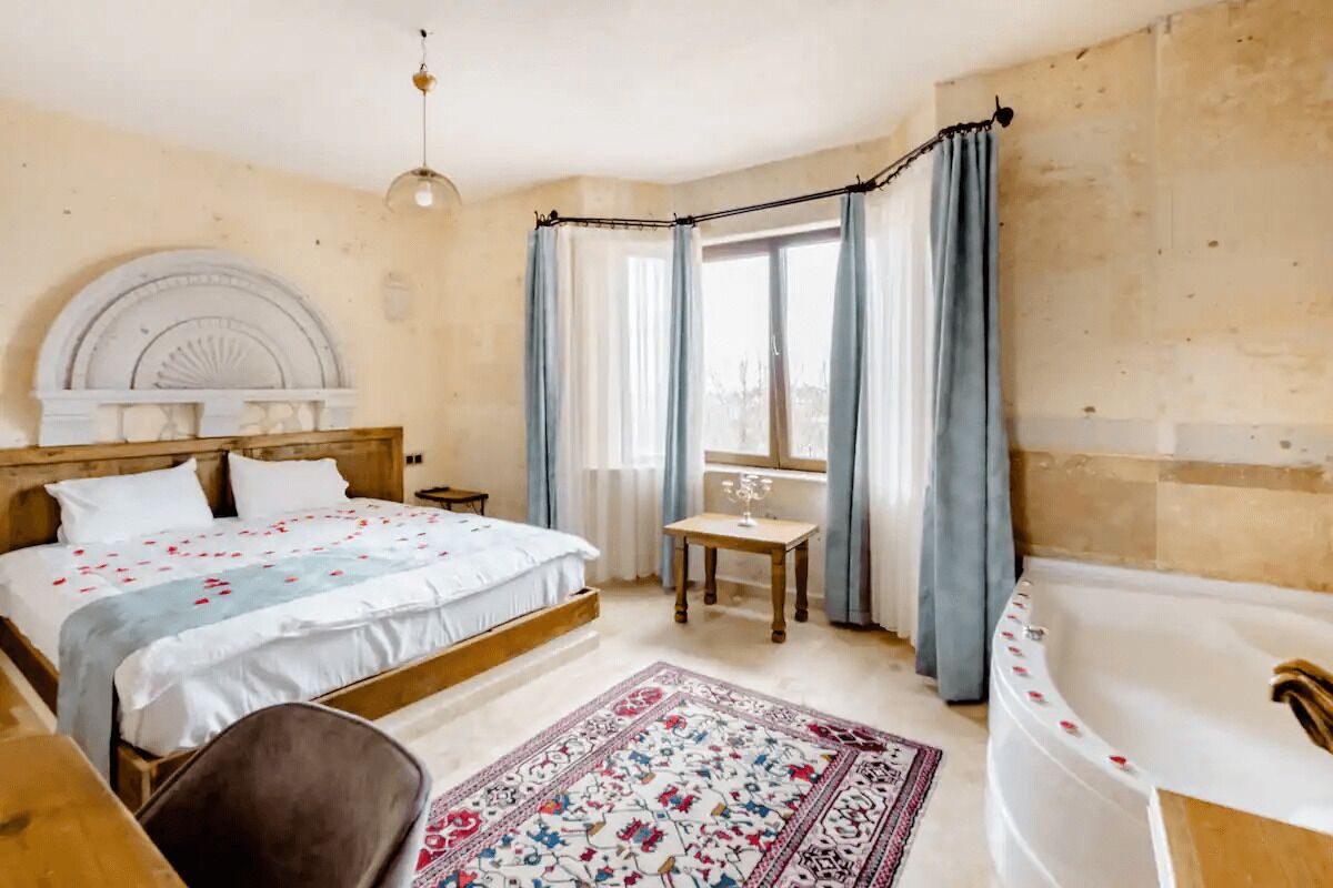 A bedroom with a large bed, a wooden headboard, a small table with a glass, a chair, a window with curtains, and a bathtub.