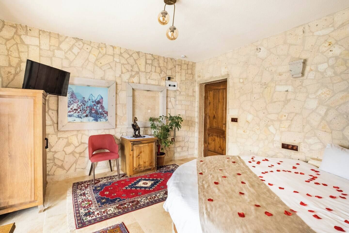 A bedroom with a stone wall, a bed with rose petals, a red chair, a wooden cabinet, and a painting.