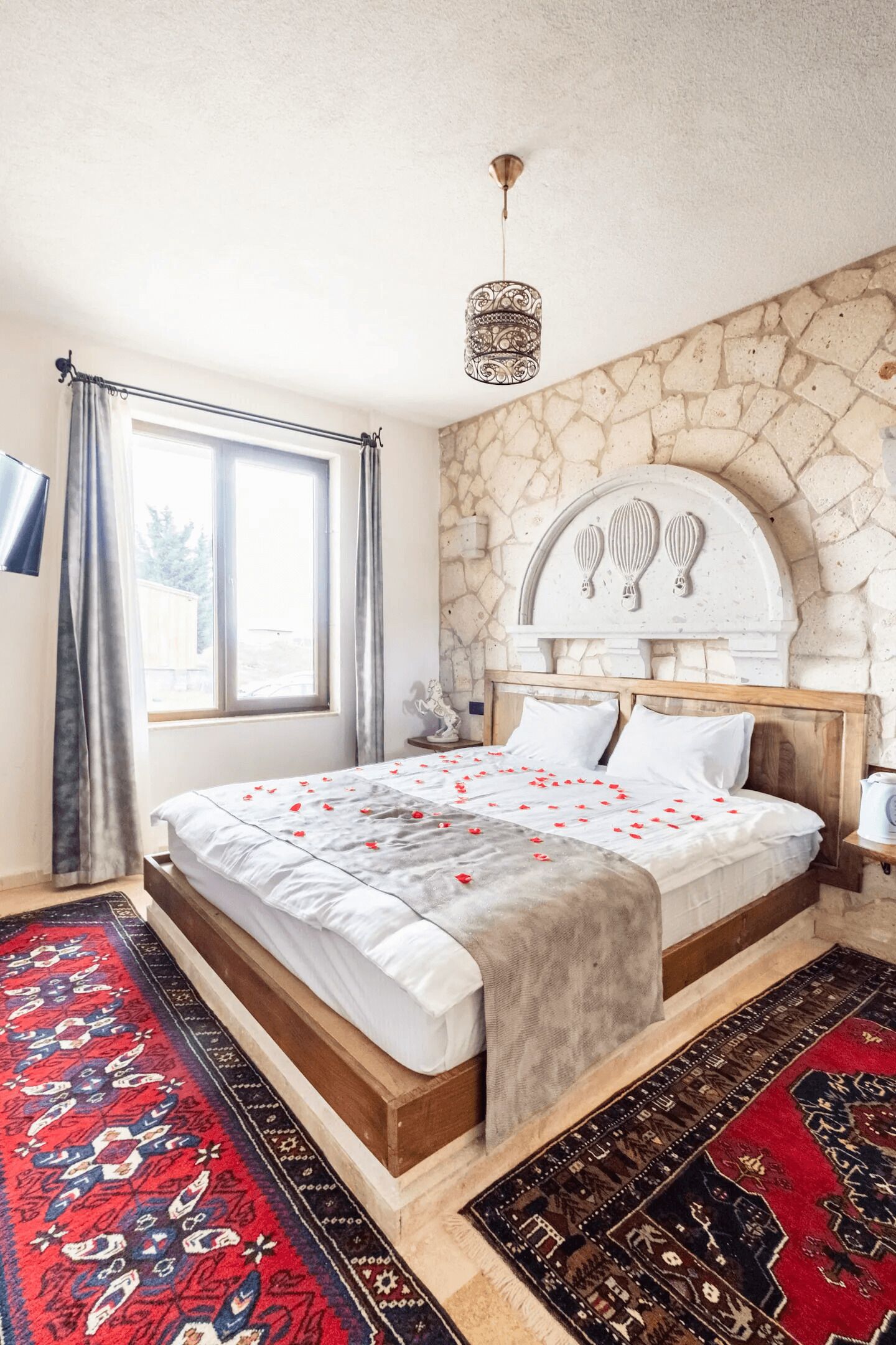 A bedroom with a stone wall, a bed with a white headboard, a red and black rug, and a window with curtains.