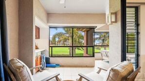 A sunlit room with a large window, two cushioned chairs, a small table with decorative items, and a view of a golf course. - Summer Deal Waikoloa Condo Near Beaches with Golf Course Views (Waikoloa)