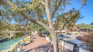 A deck with outdoor seating overlooking a river, a parking area, and a building with a stone facade. - IC 209 Comal Escape (NEW BRAUNFELS)