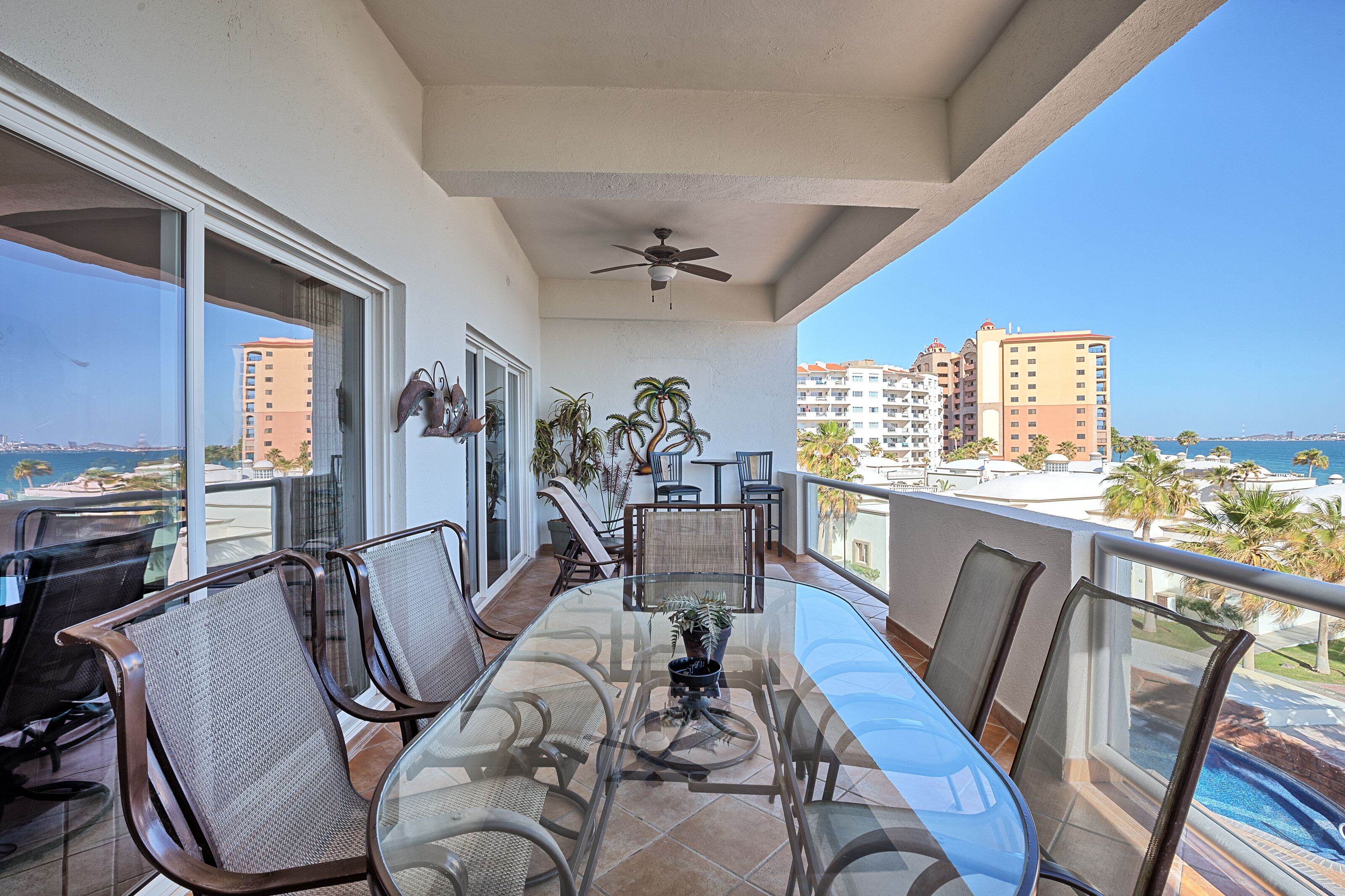 A balcony with a glass table and chairs, a ceiling fan, and a view of the ocean and buildings.