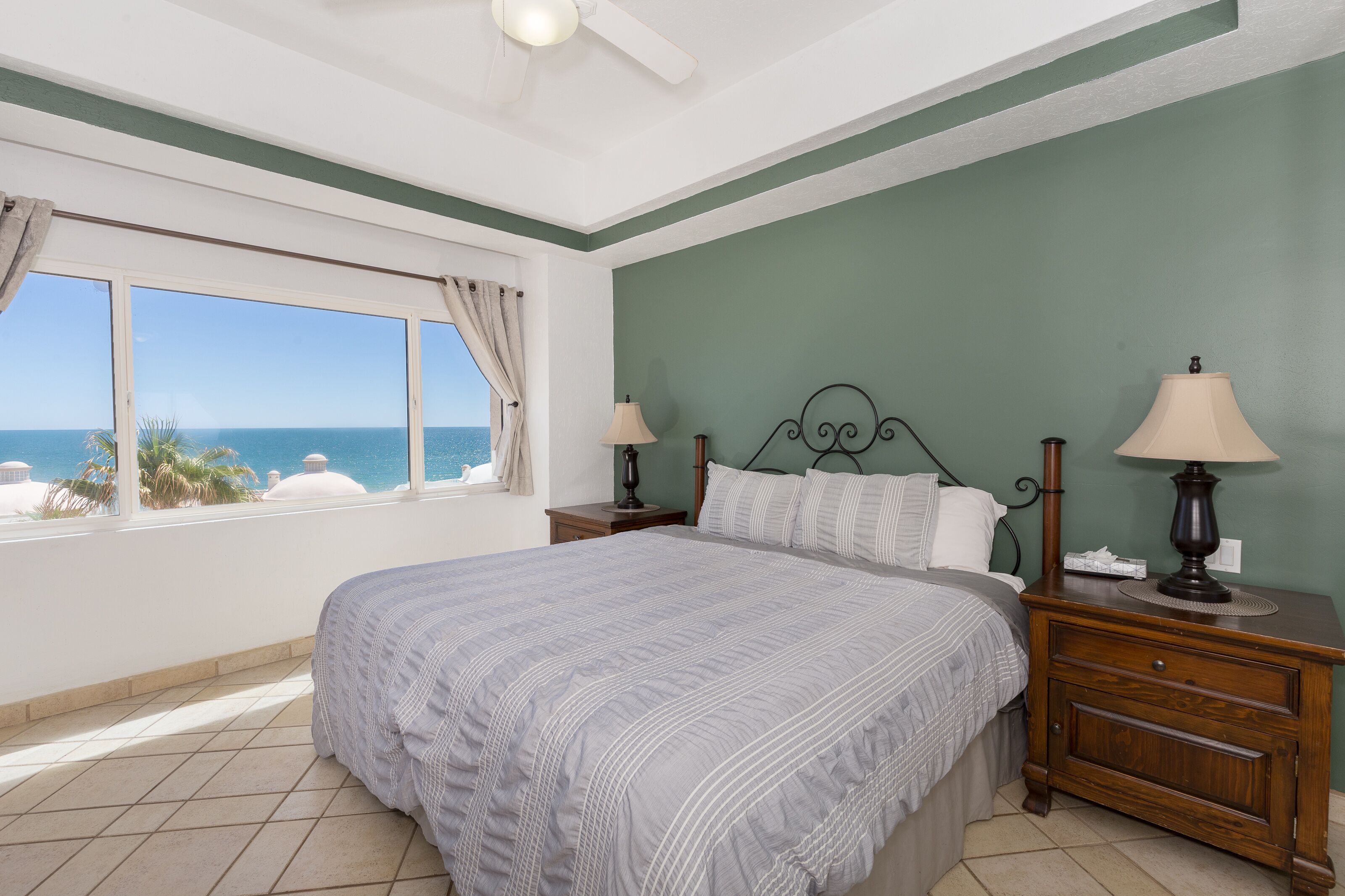 A bedroom with a large bed, two bedside tables with lamps, a view of the ocean, and a ceiling fan.