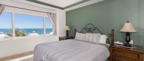 A bedroom with a large bed, two bedside tables with lamps, a view of the ocean, and a ceiling fan.