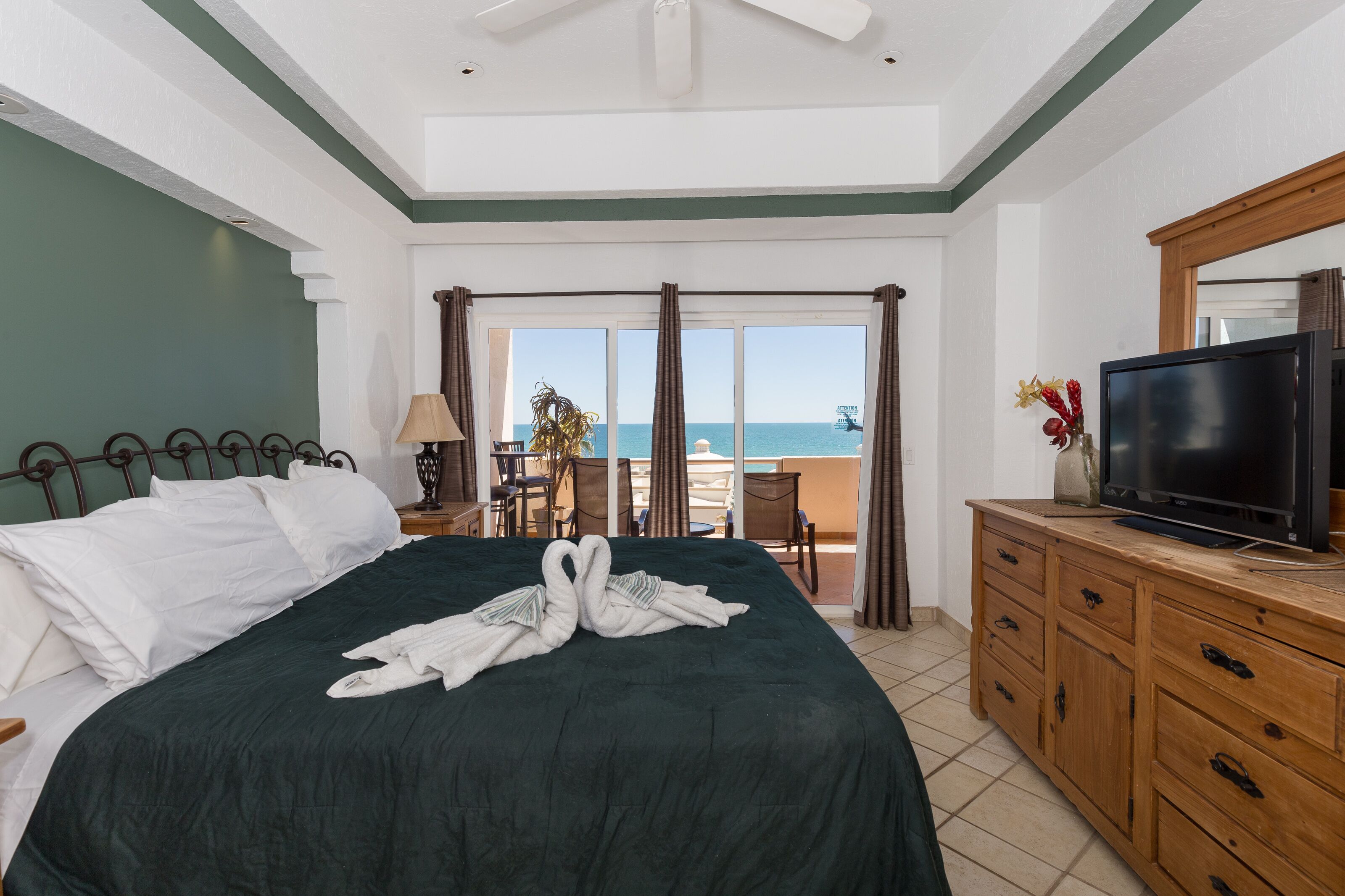 Habitación de hotel con cama, televisión, cómoda de madera y balcón con vista al mar.