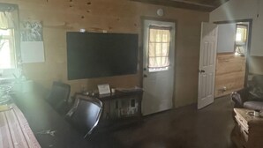 A spacious room with a large flat-screen TV, a wooden ceiling, a door with a window, and a small table with chairs. - Quiet cabin in the hills close to the trails and other outdoor activities.<br> (Genoa)