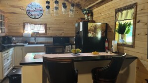 A cozy kitchen with wooden walls, a large clock, hanging pendant lights, a refrigerator with a fruit painting, and a bar area with stools. - Quiet cabin in the hills close to the trails and other outdoor activities.<br> (Genoa)