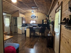 Interior acogedor de una cabaña, con revestimiento de madera, una zona de cocina con fregadero y encimera, y un comedor con banco y sillas.