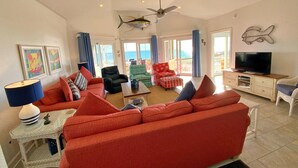 A spacious living room with a large red sofa, a black armchair, and a green patterned ottoman. There is a ceiling fan, a flat-screen TV on a wooden cabinet, and a small white table with a lamp. - Beachfront, Pools, Pickleball, in Morgantown (Gulf Shores)
