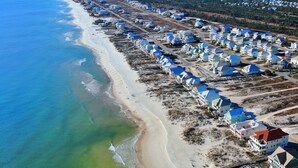 An aerial view of a coastal residential area with houses aligned along the beach, a clear view of the ocean, and a distant shoreline. - Beachfront, Pools, Pickleball, in Morgantown (Gulf Shores)