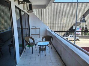 A balcony with wicker chairs and a small table, overlooking a street with a parked car and a building with vertical siding.