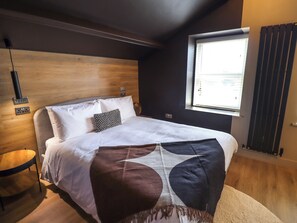 A modern bedroom with a wooden headboard, a large bed with white linens, a patterned throw pillow, a round side table, and a window with blinds. - Glencoe House (Wooler)